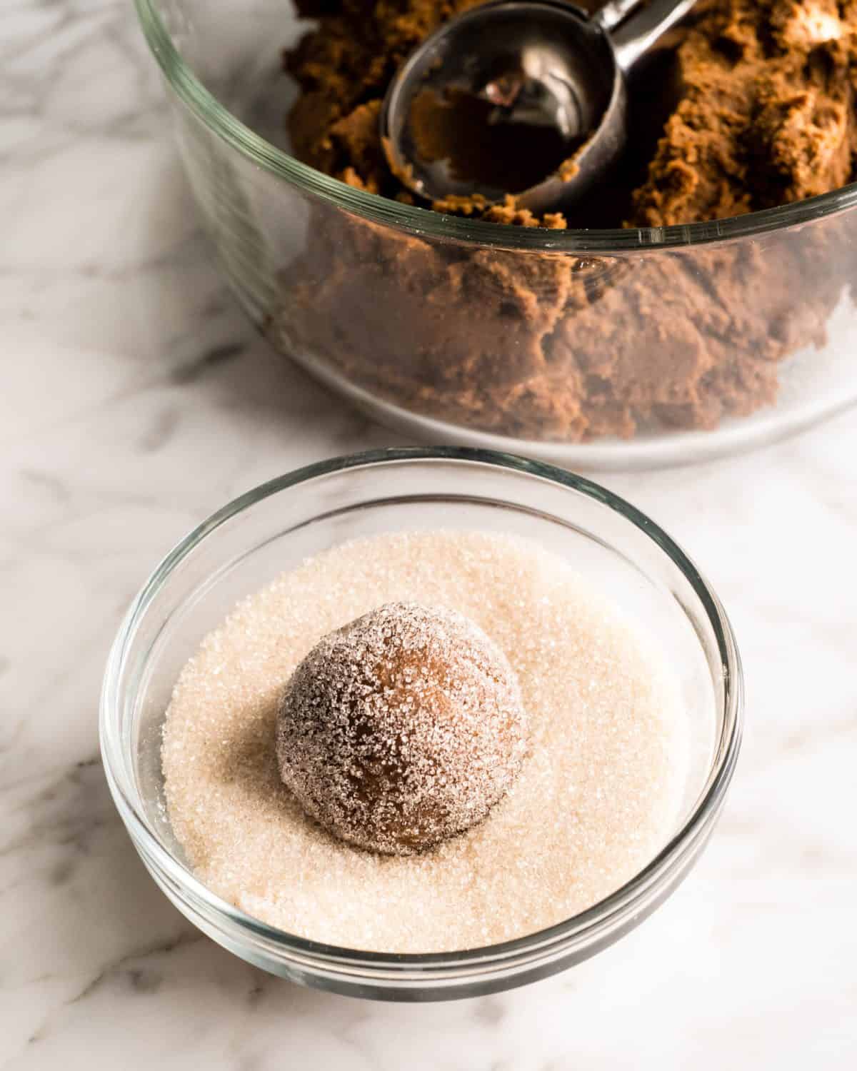 a ball of soft gingerbread cookies dough in a small bowl of granulated sugar - it has been rolled in the sugar so it's coated. The glass bowl of dough with a cookie scoop is in the background. Everything is on a marble surface.