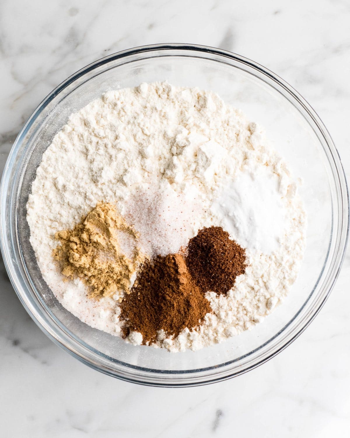 making soft gingerbread cookies - flour, salt, baking soda, cinnamon, ginger, and cloves in a glass bowl on a marble surface before mixing