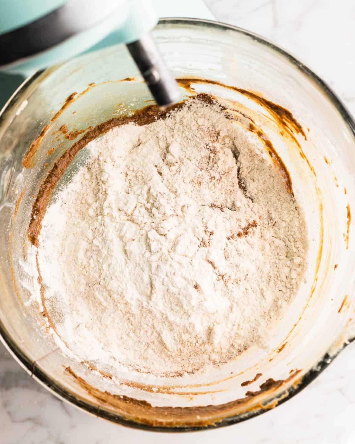 making easy gingerbread cookies - flour mixture added to the wet ingredients in a glass mixing bowl of a light blue standing mixer before beating together