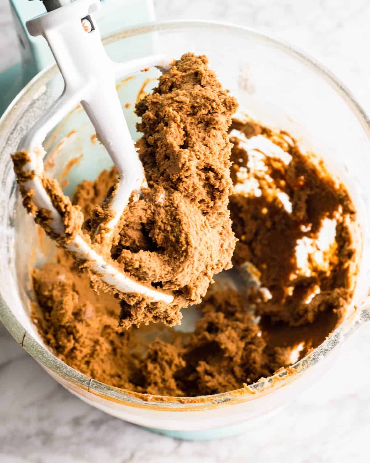 final easy gingerbread cookies dough in a glass bowl of a light blue standing mixer fitted with the paddle attachment, there is dough on the attachment and in the bowl.