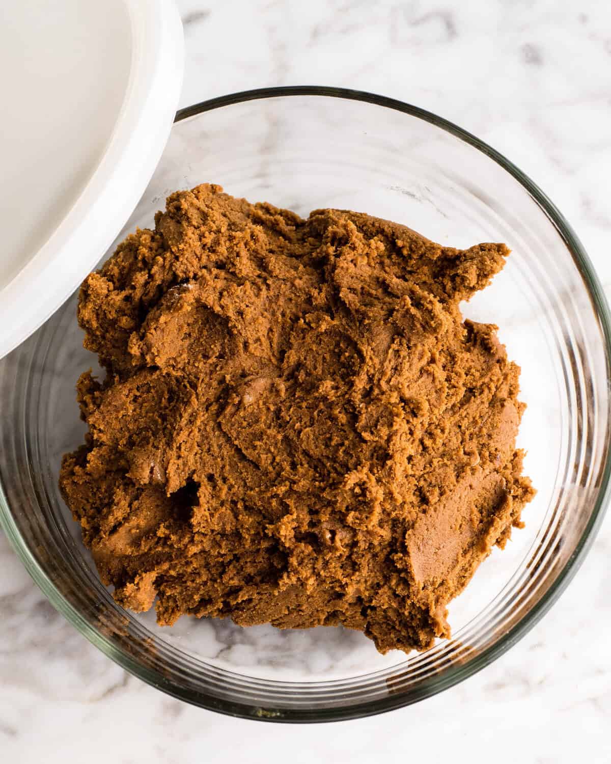 These Soft Gingerbread Cookies are the BEST gingerbread cookies ever! They have crispy edges with soft and chewy centers and are perfectly spiced! Plus they are so easy to make - no rolling or cutting-out necessary to enjoy this rich & nostalgic gingerbread recipe! Roll them in sugar, add a simple gingerbread glaze or serve them plain! #gingerbreadcookies #christmascookies #gingerbread soft gingerbread cookies dough in a glass bowl with a white lid on a marble surface to be chilled.