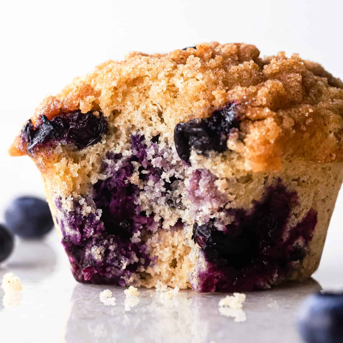 one blueberry muffin with a bite taken out of it so the inside crumb and blueberries can be seen, with blueberries and small crumbs around it