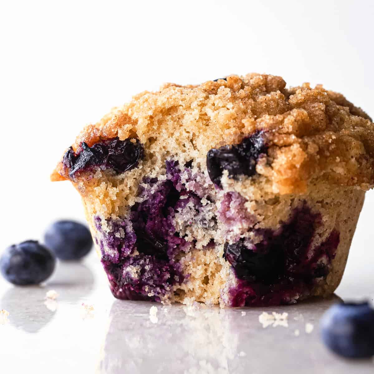 one blueberry muffin with a bite taken out of it so the inside crumb and blueberries can be seen, with blueberries and small crumbs around it
