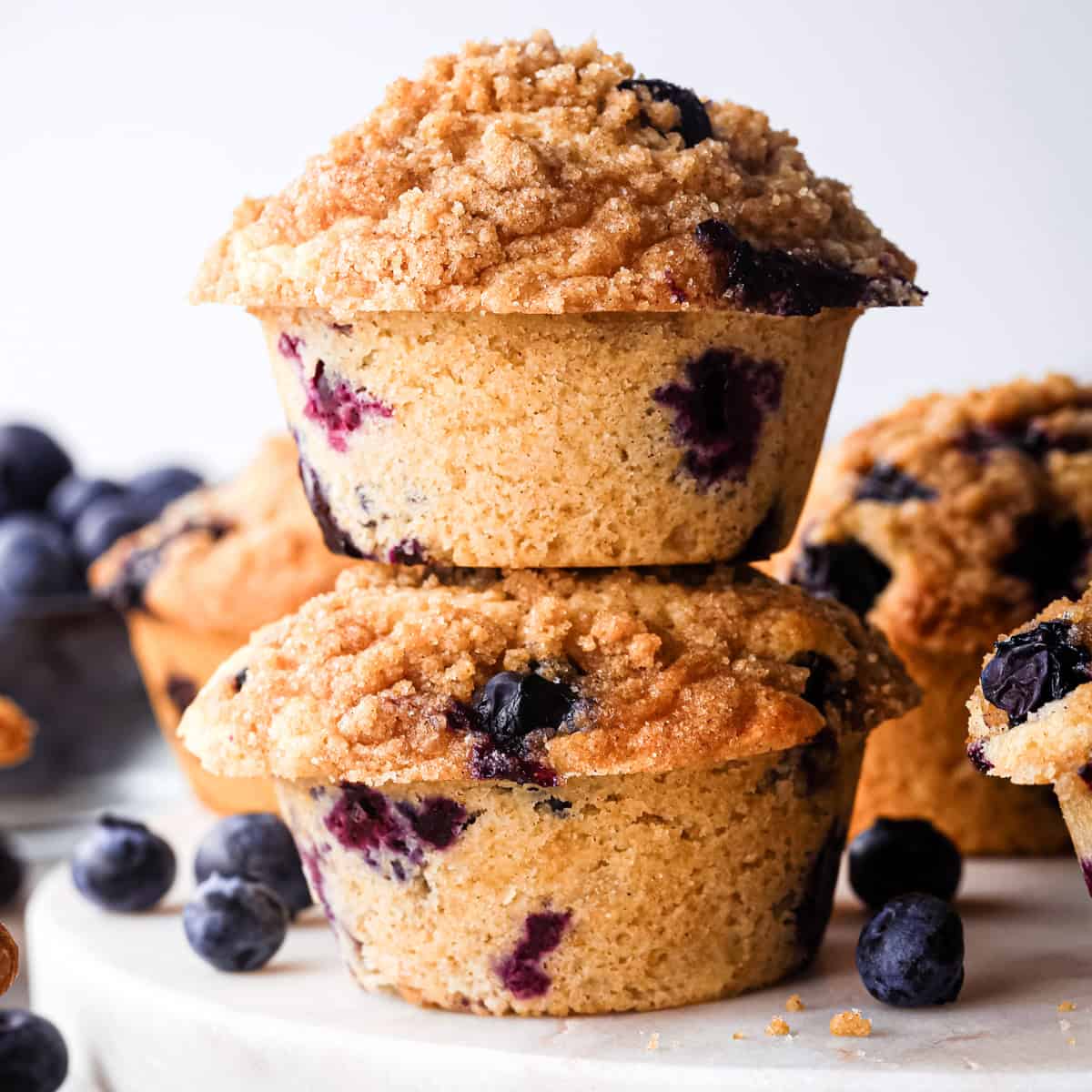 a stack of two blueberry muffins with crumb topping, surrounded by blueberries and 3 additional blueberry muffins