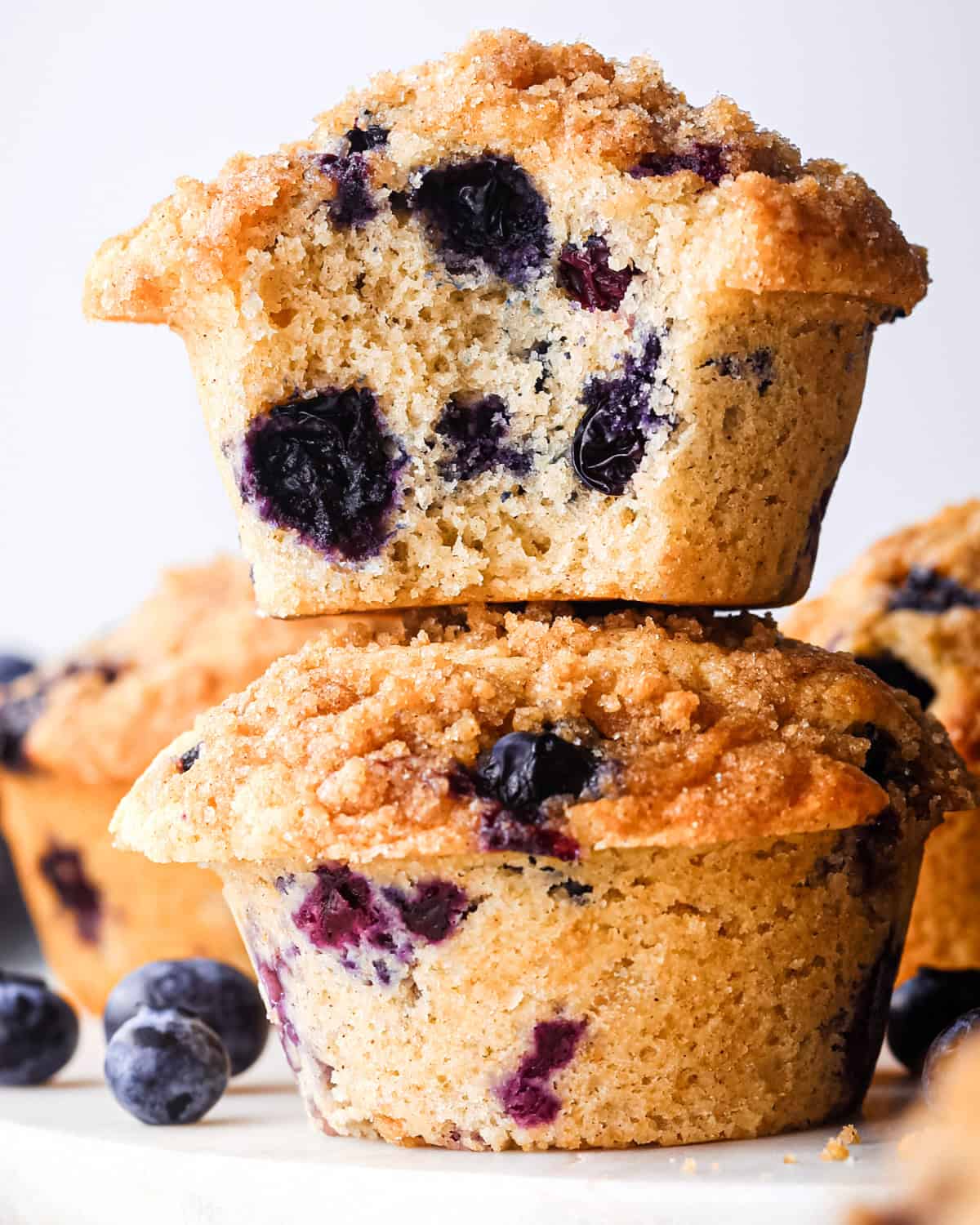 a stack of two blueberry muffins with crumb topping, the top one has a bite taken out fo it so the inside crumb and blueberries can be seen, surrounded by blueberries and  additional blueberry muffins