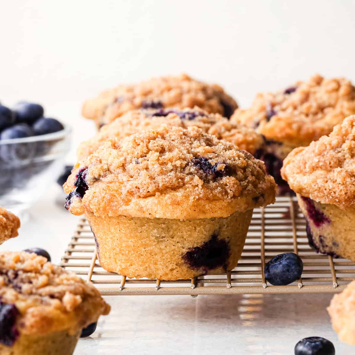 5 blueberry muffins on a wire cooling rack with crumb topping surrounded by blueberries and 3 other muffins. 