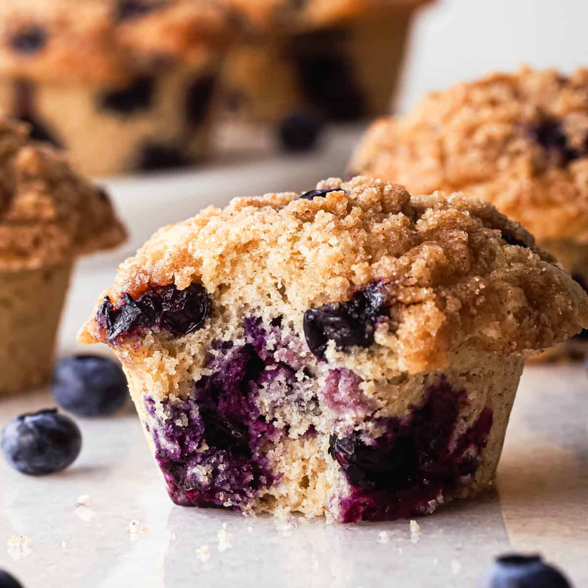a blueberry muffin with a bite taken out of it so the inside crumb and blueberries can be seen, with blueberries and 4 other muffins around it