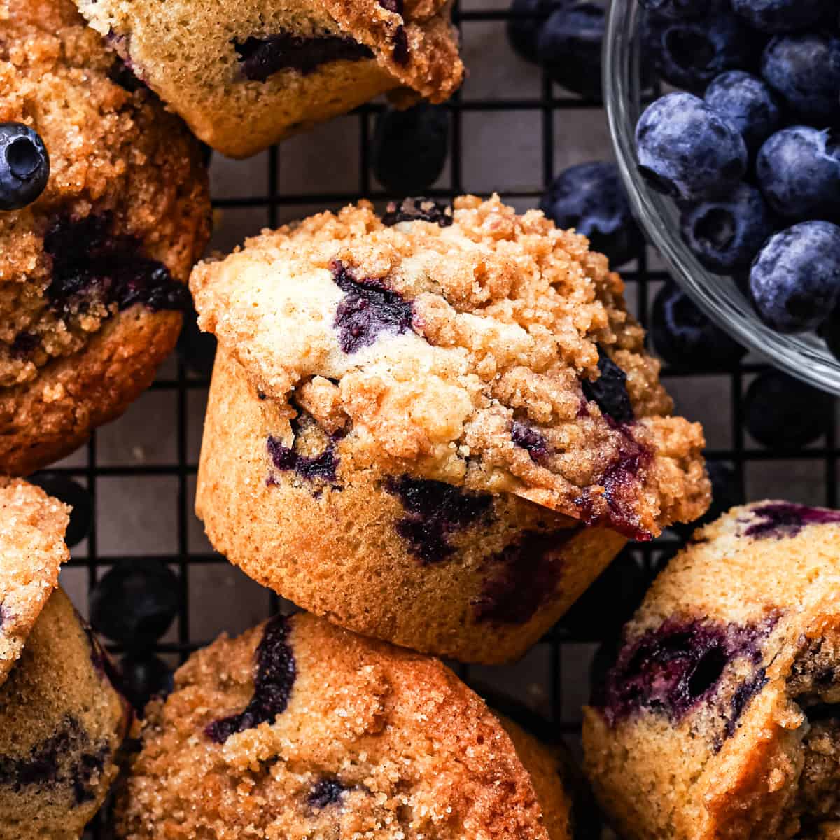 six blueberry muffins with crumb topping on a wire cooling rack surrounded by blueberries
