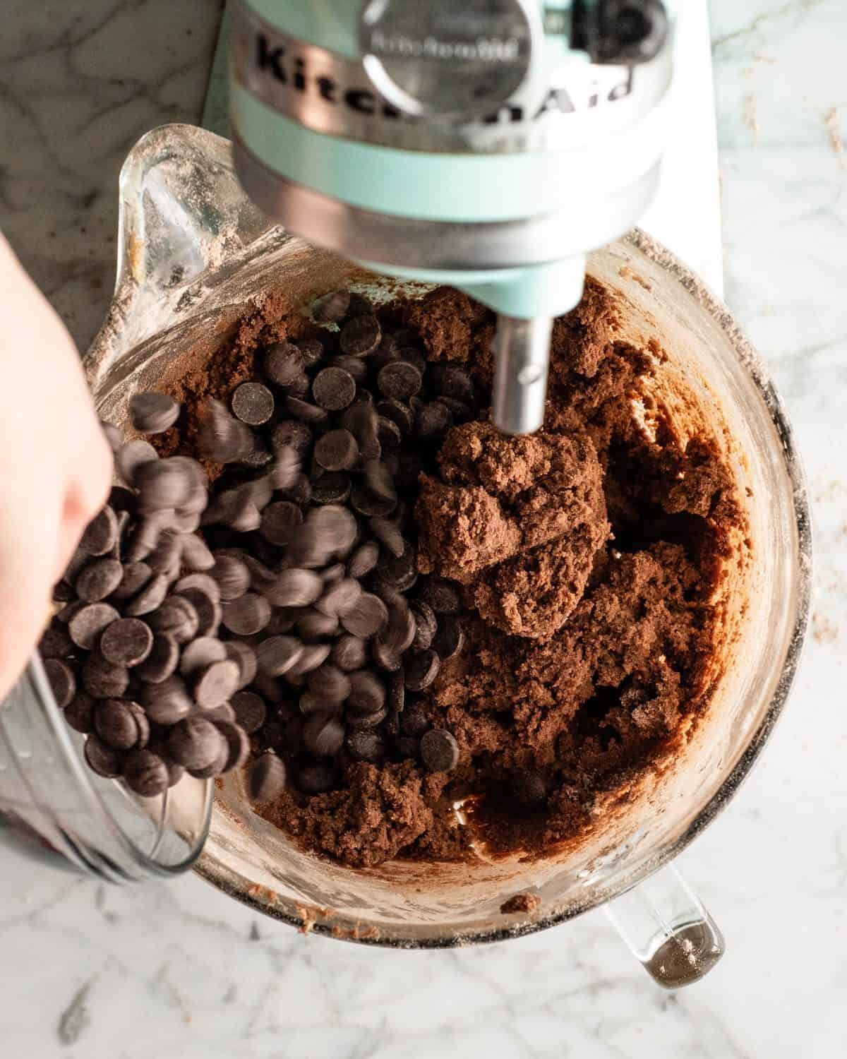 making double chocolate chip cookies - chocolate chips being poured into the glass mixing bowl of a light blue standing mixer with the chocolate cookie dough in it