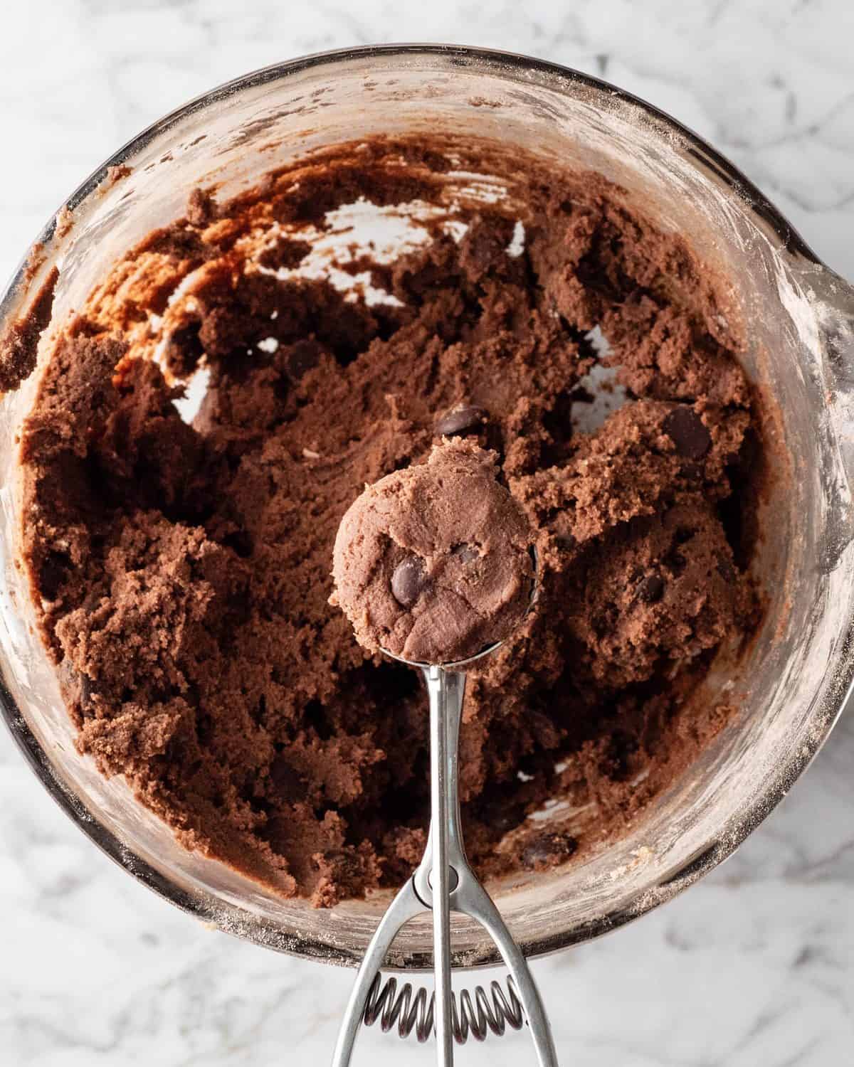 making double chocolate chip cookies - a cookie scoop with dough in it being held over the glass bowl with the rest of the chocolate cookies dough in it on a marble surface