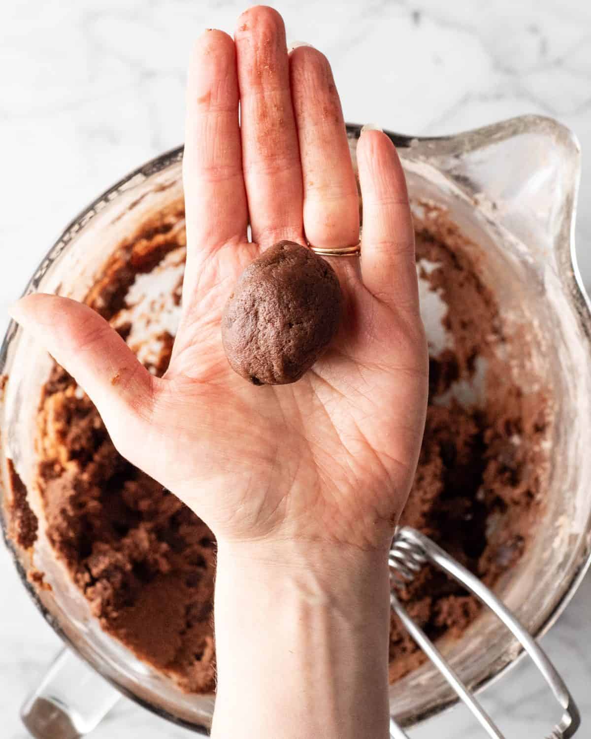 making double chocolate chip cookies - a hand holding a cookie dough ball over the bowl of cookie dough on a marble surface