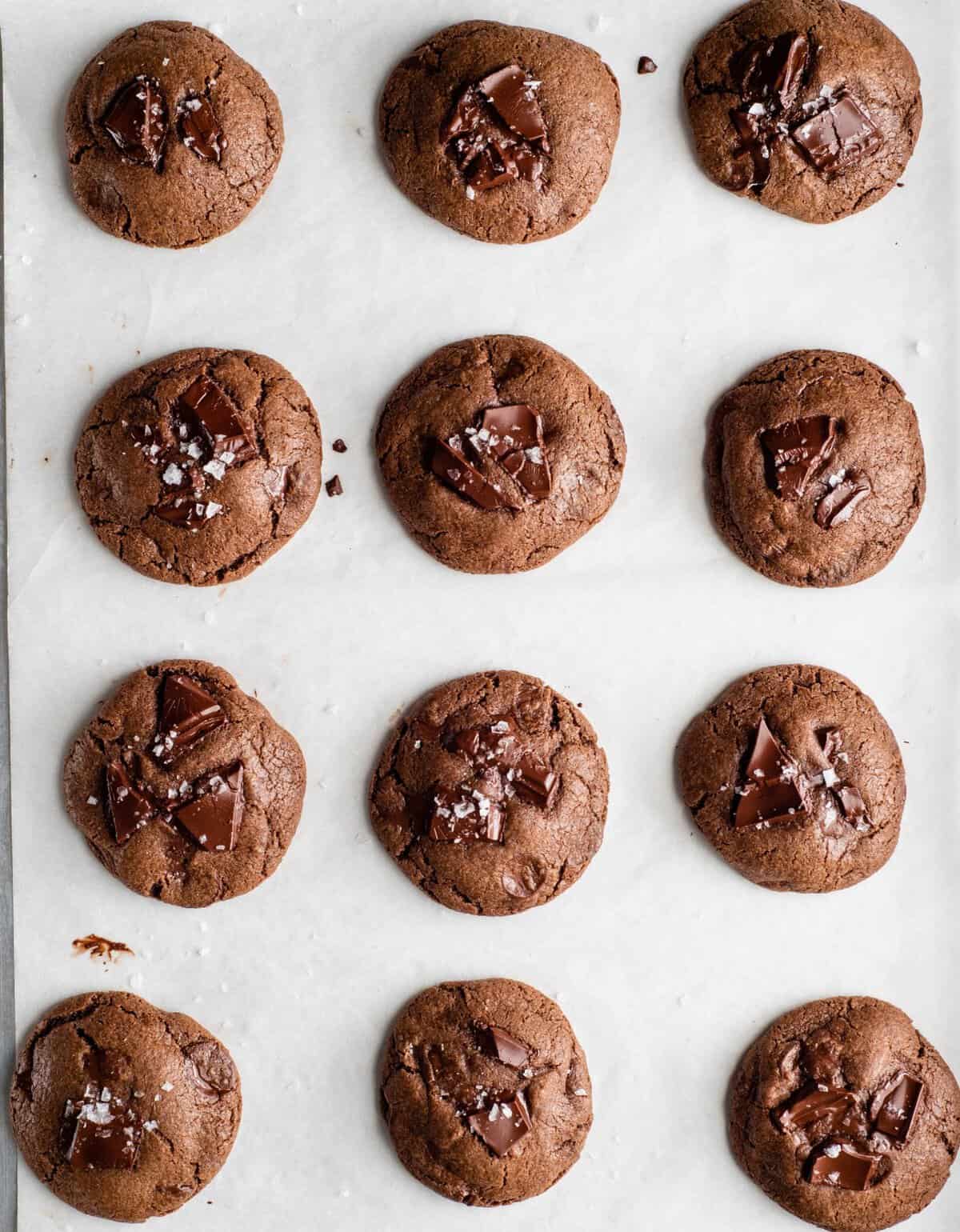 overhead photo of 12 baked double chocolate chip cookies on a baking sheet lined with parchment paper they all have melty chopped chocolate on top and flaky sea salt
