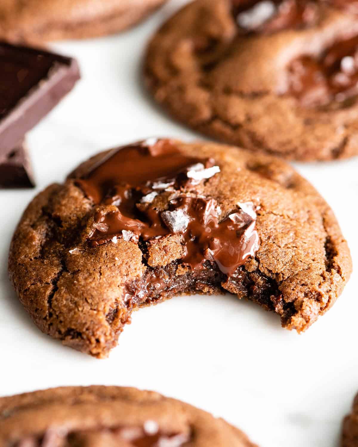 The BEST easy double chocolate chip cookie recipe ever! These chocolate cookies have soft centers, chewy edges and are irresistibly chocolatey. They come together in 5 minutes and require only 30 minutes of chilling time! #chocolate #cookies #chocolatecookies #baking #joyfoodsunshine 3 double chocolate chip cookies with melty chopped chocolate and flaky sea salt on top all on a marble surface. The center cookie has a bite taken out of it so the gooey fudgy texture is visible, there's a stack of chocolate in the background