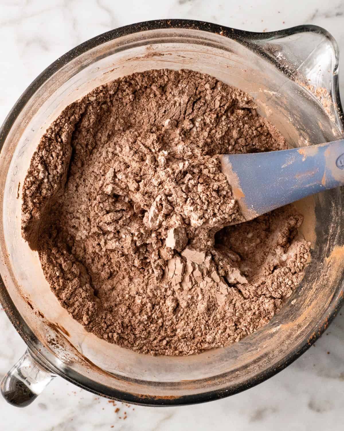 making double chocolate chip cookies -dry ingredient mixture including cocoa powder in a glass batter bowl being mixed with a blue spatula