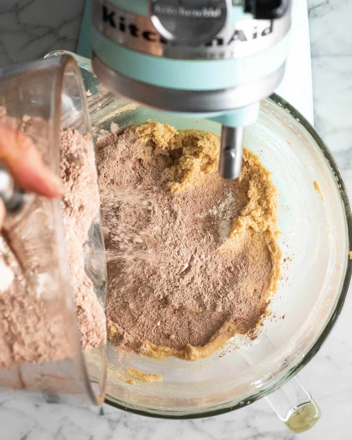 making double chocolate chip cookies - dry ingredient mixture being poured from a glass batter bowl into the glass mixing bowl of a light blue standing mixer with the beaten wet ingredients in it