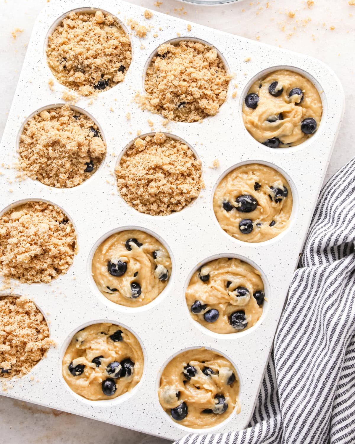 blueberry  muffins batter in a muffin pan before baking, 6 muffins have crumb topping on them, and there is a white and blue striped towel nearby. 