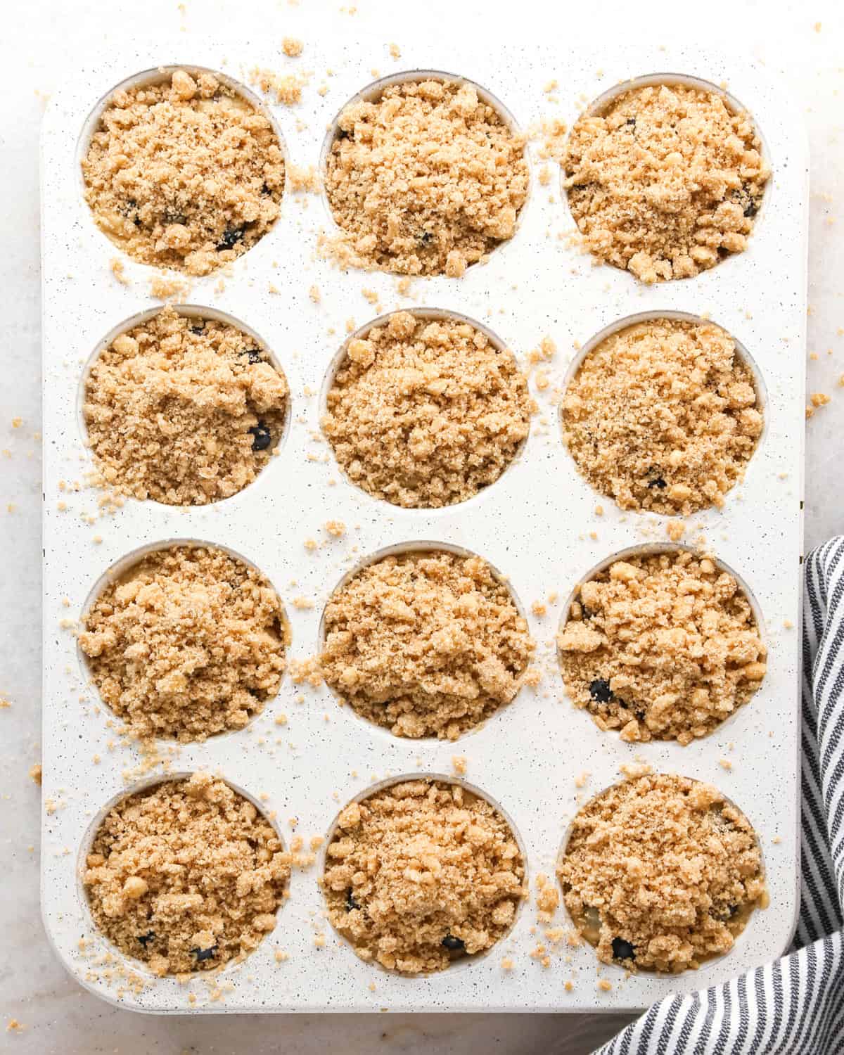 blueberry  muffins batter in a muffin pan before baking with crumb topping on top and a striped towel nearby