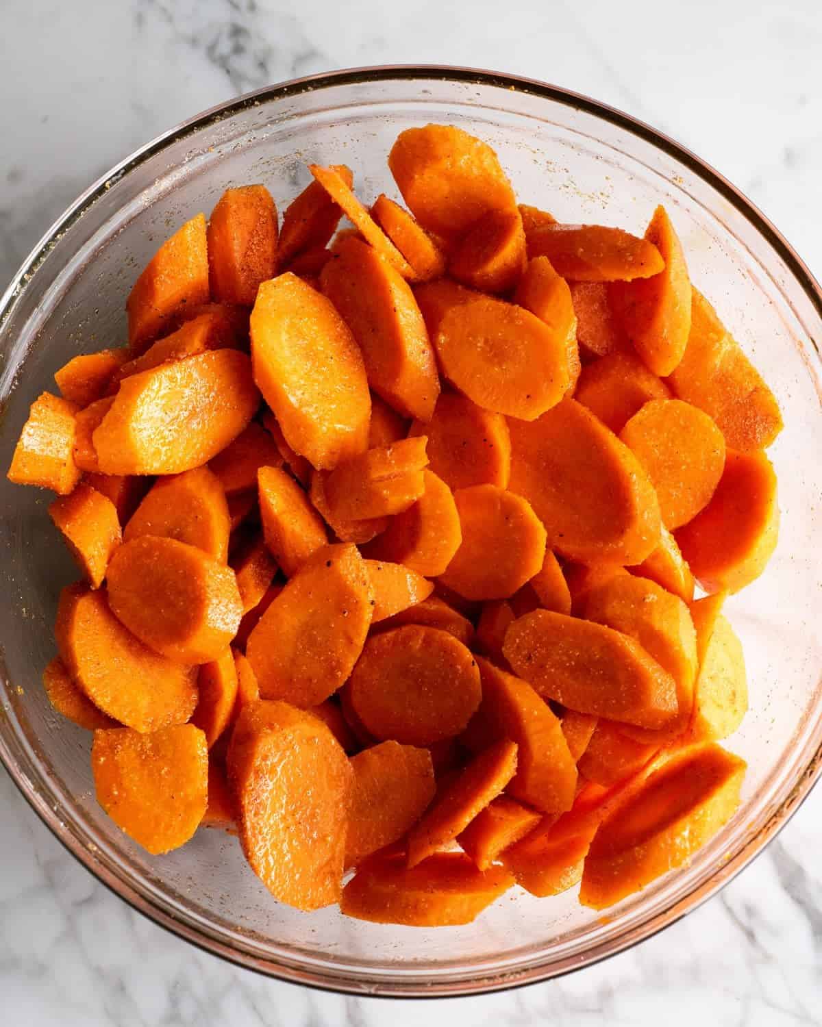 how to make oven roasted carrots recipe - oiled and seasoned carrot rounds in a glass bowl on a marble surface