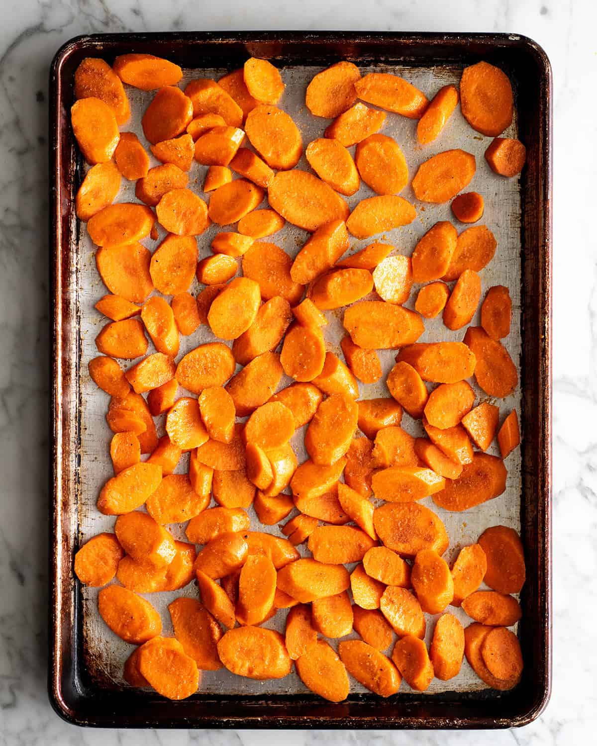 how to make oven roasted carrots recipe - oiled and seasoned carrot rounds spread out onto a metal baking sheet on a marble surface before roasting