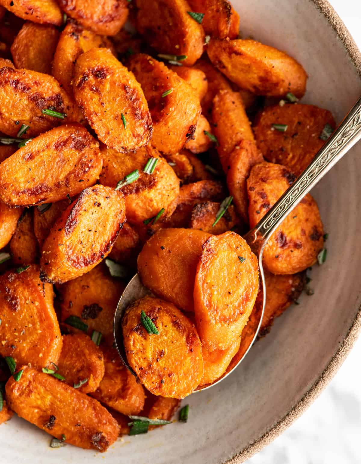 oven roasted carrots cut into carrot rounds in a white ceramic bowl. The outside is golden brown and crispy and they are garnished with chopped rosemary and there's a silver spoon in the roasted carrots taking a scoop.