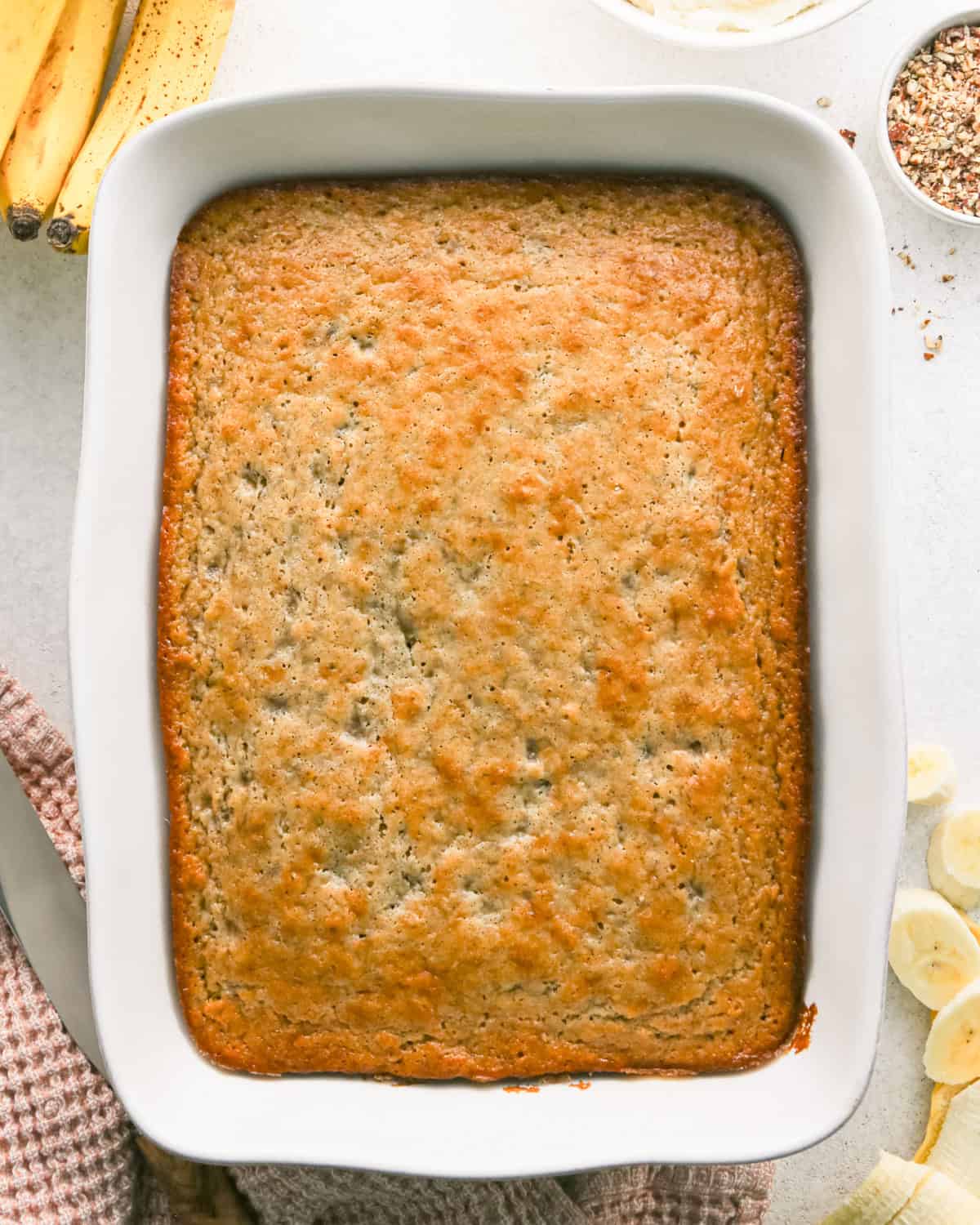 banana cake in a white baking dish after baking, with bananas and chopped nuts around the baking pan
