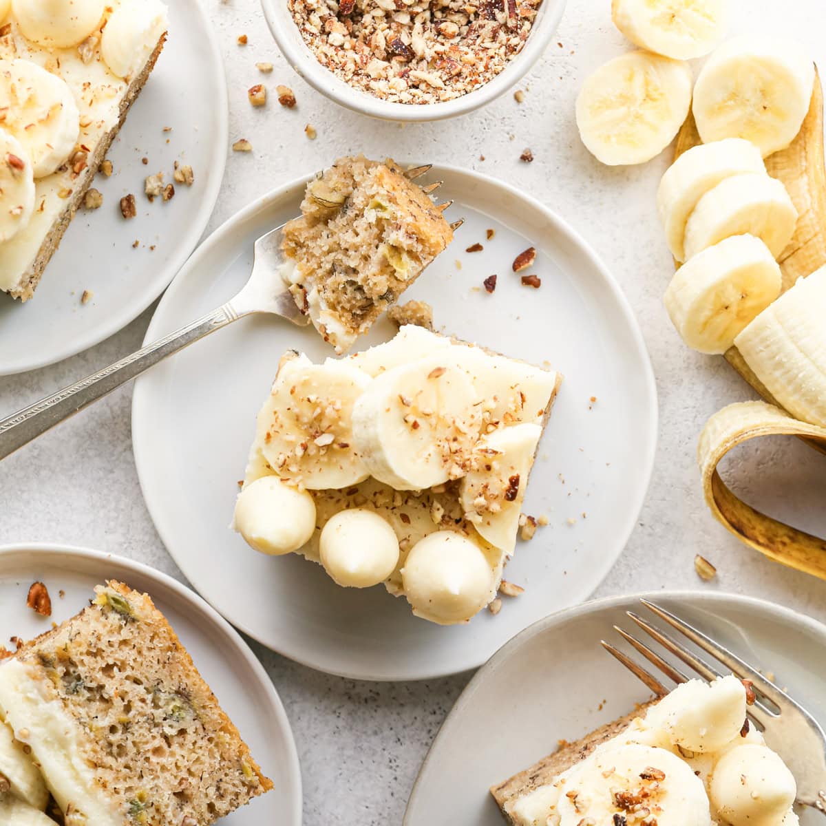 4 plates, each with a slice of banana cake on it with cream cheese frosting. The middle piece has a fork with a bite of banana cake on it. all surrounded by chopped nuts and banana slices 