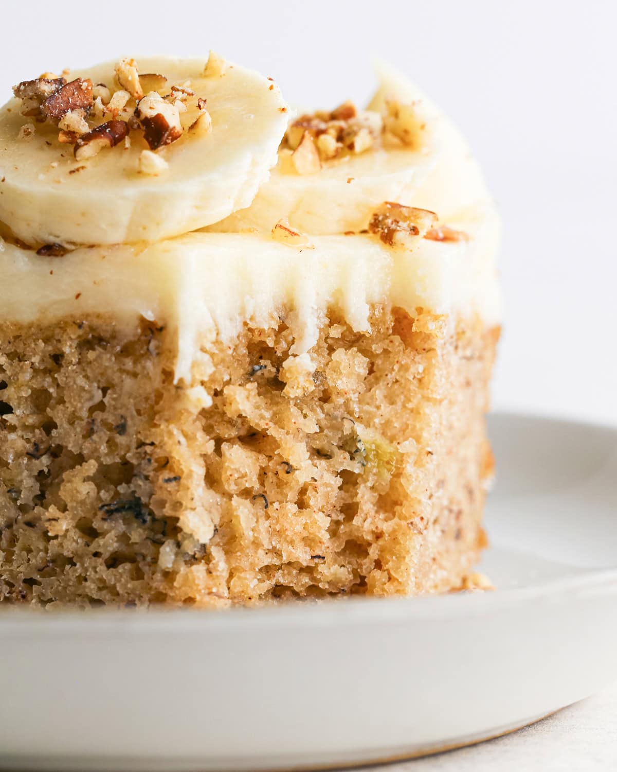 a close up photo of a piece of banana cake with a bite taken out of it, topped with cream cheese frosting, banana slices, and a sprinkle of chopped nuts