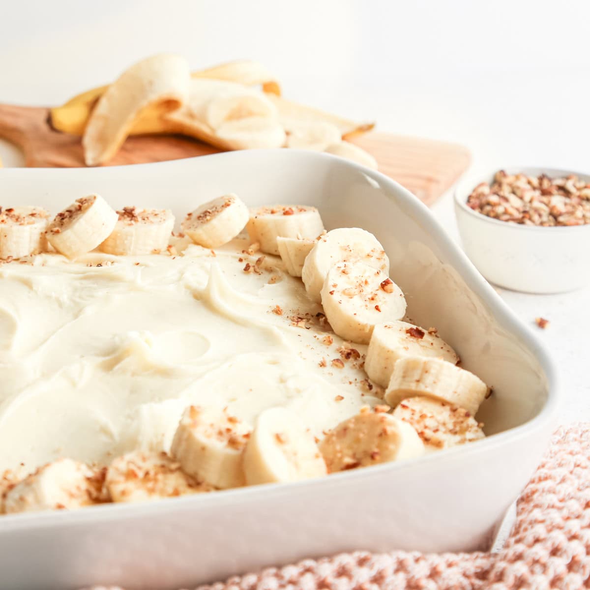 front view of a banana cake with cream cheese frosting toppd with banana slices and chopped nuts