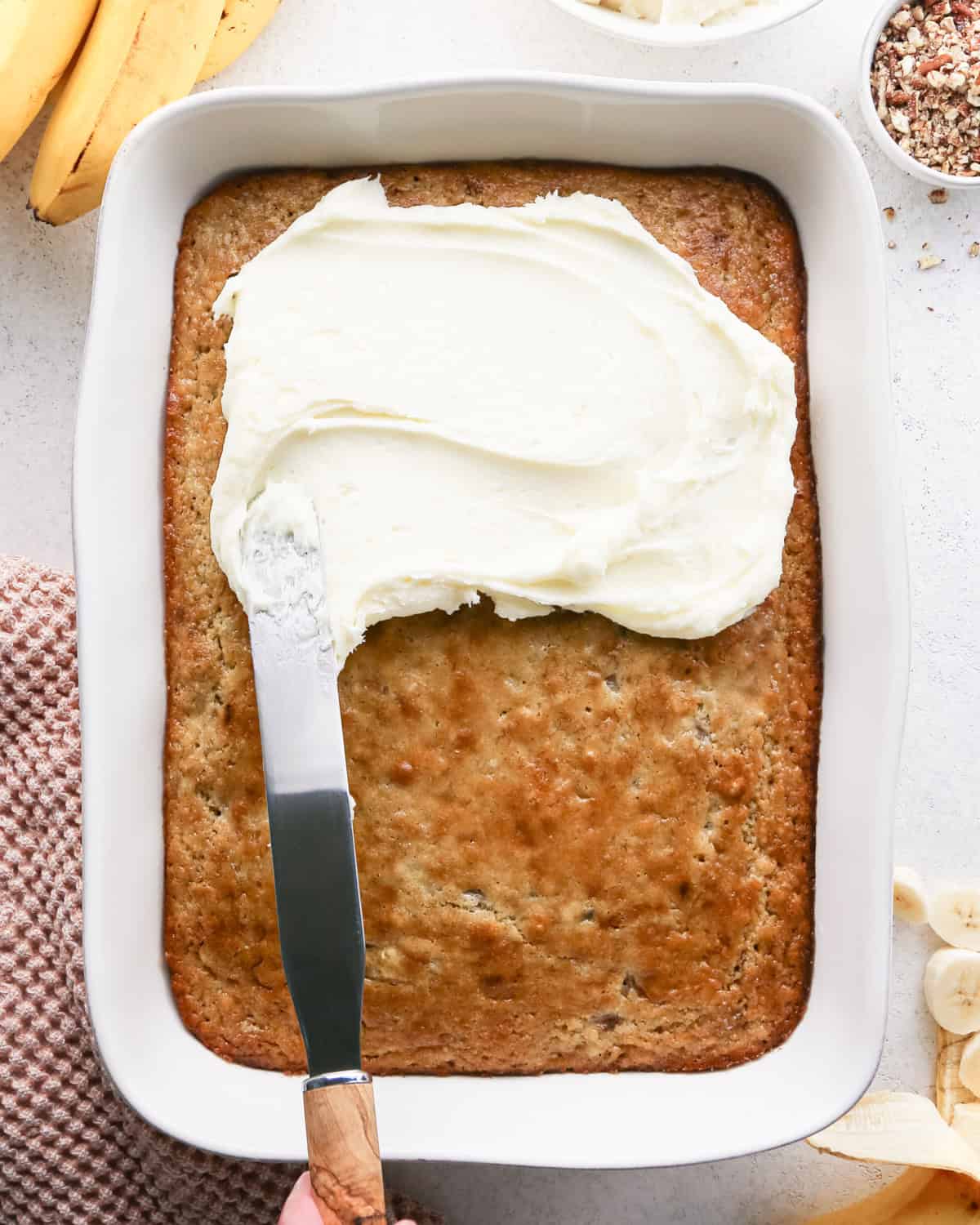 cream cheese frosting being spread on top of banana cake in a baking dish with a metal spatula