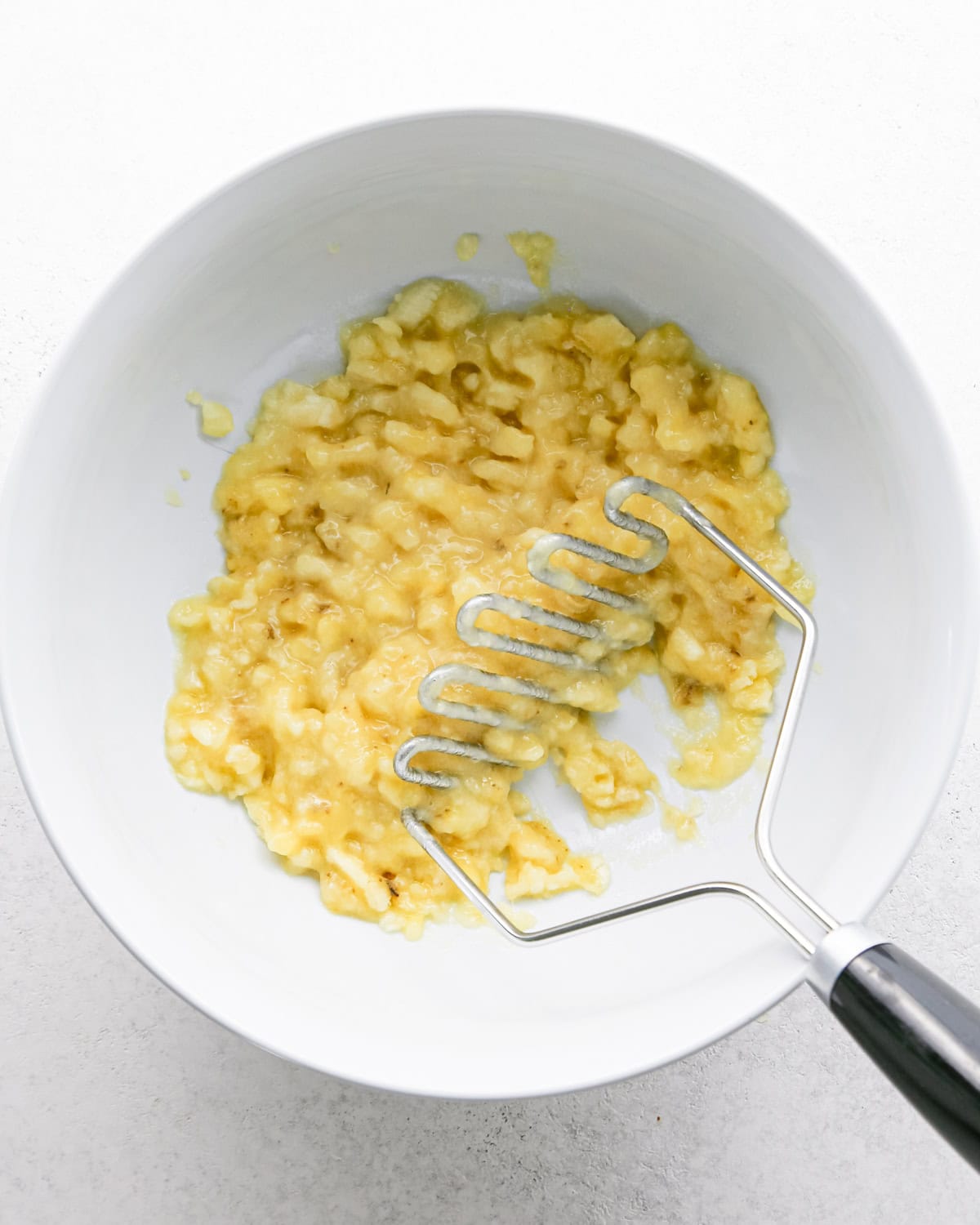 mashed banana in a white bowl with a banana masher to make banana cake