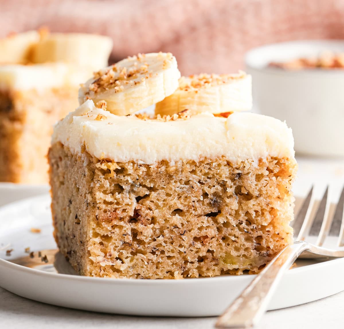 a moist slice of banana cake topped with cream cheese frosting, banana slices, and a sprinkle of chopped nuts, served on a white plate with a fork.