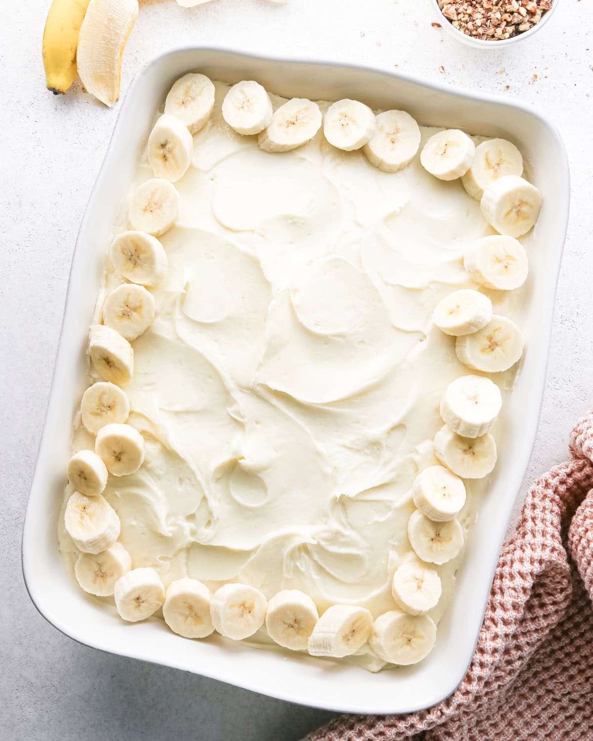 sliced bananas arranged around the outside edge of a banana cake with cream cheese frosting in a white baking dish