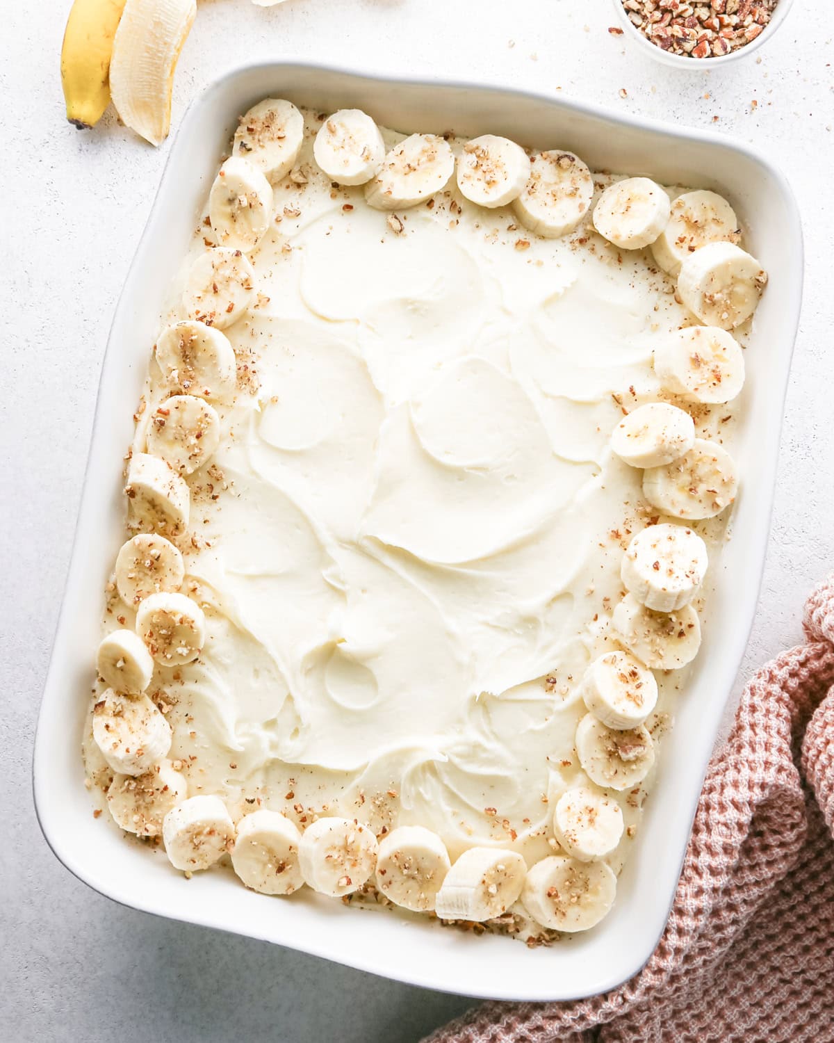 chopped nuts sprinkled over the sliced bananas arranged around the outside edge of a banana cake with cream cheese frosting in a white baking dish