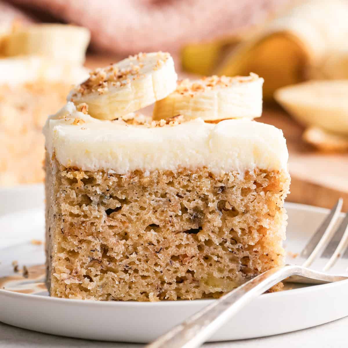 This is the best banana cake recipe ever! It's easy to make (no mixer required), moist, loaded with bananas and topped with a delicious cream cheese frosting. a moist slice of banana cake topped with cream cheese frosting, banana slices, and a sprinkle of chopped nuts, served on a white plate with a fork.