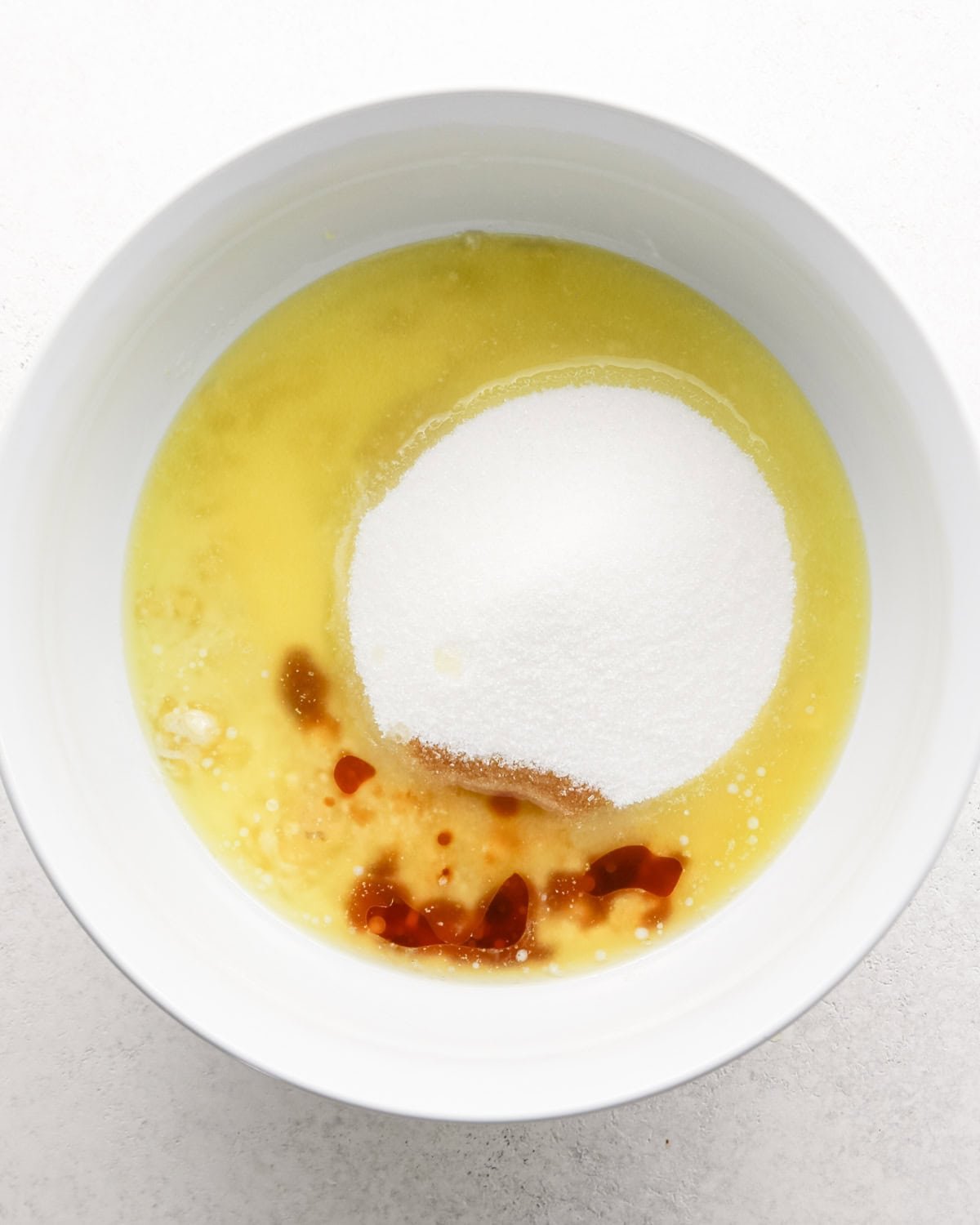 a white bowl with sugar, vanilla and melted butter added to the mashed banana before mixing to make banana cake