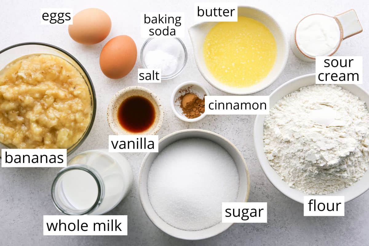 overhead photo of the ingredients in this Banana Cake Recipe like eggs, mashed banana, milk, vanilla, sugar, flour, melted butter, sour cream, cinnamon baking soda and salt