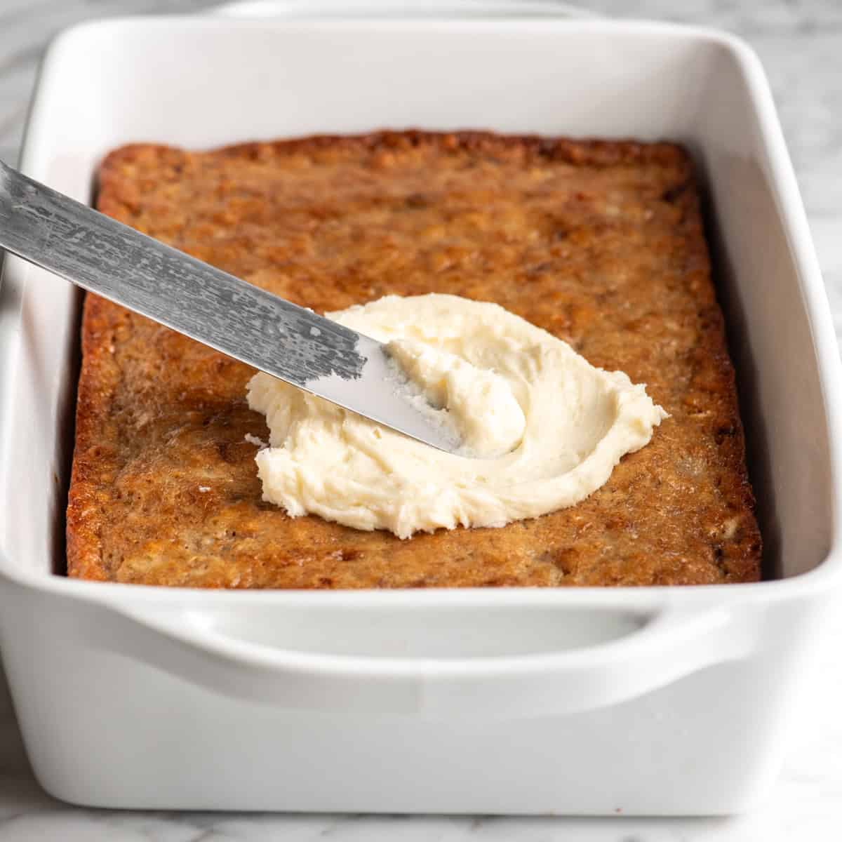 a metal spatula spreading cream cheese frosting on top of a banana cake in a white ceramic baking dish