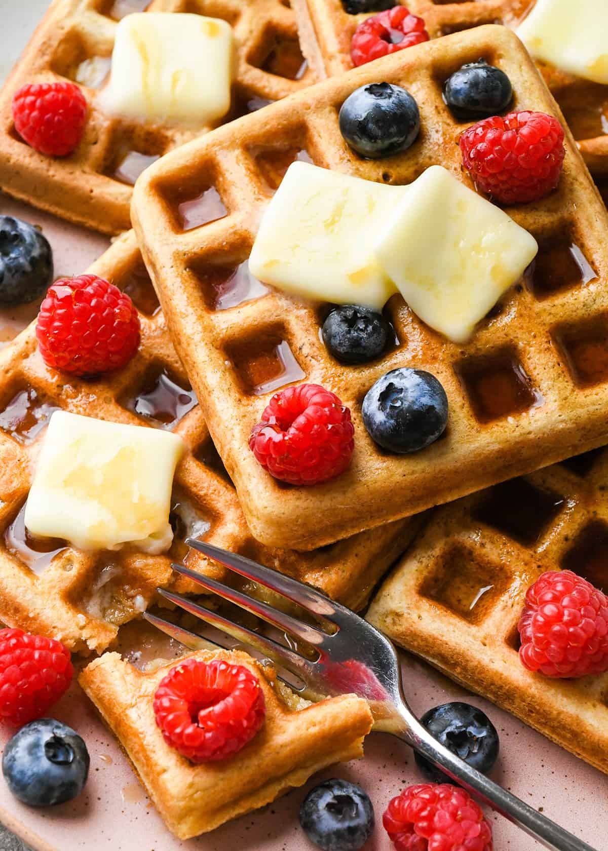 6 waffles on a plate with syrup, butter and berries with a fork taking a bite of one waffle