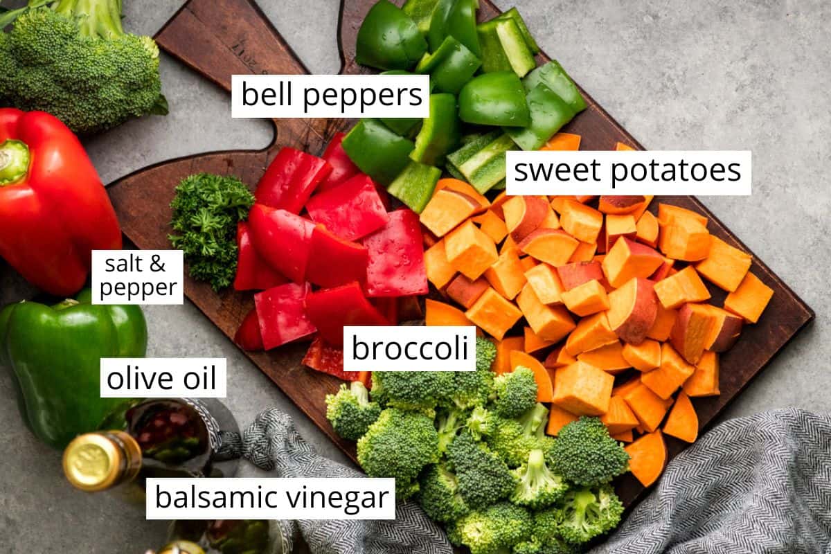 Overhead view of raw, cut up broccoli, sweet potatoes, red & green bell peppers on a cutting board surrounded by bottles of olive oil and balsamic vinegar 