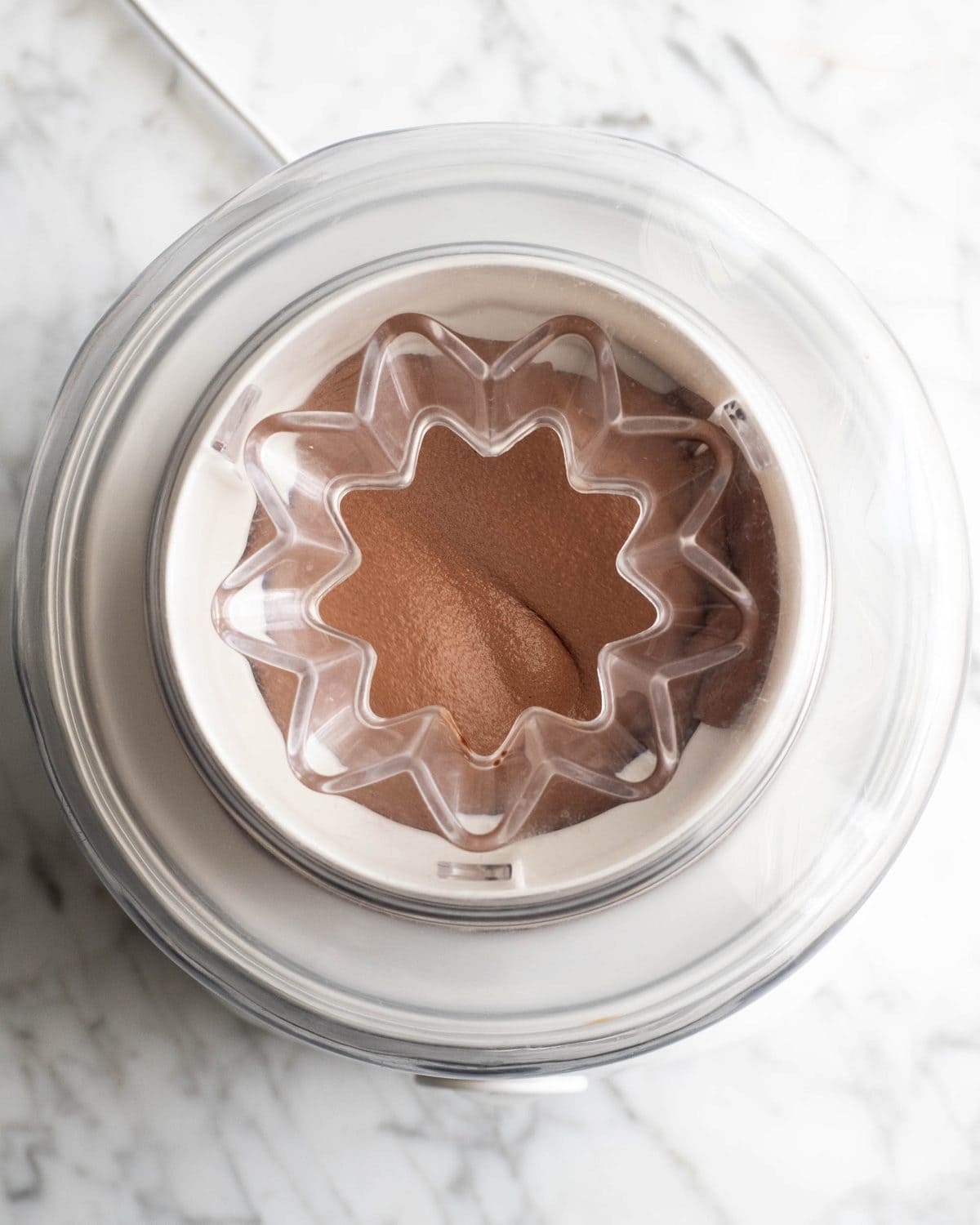 how to make chocolate ice cream - chocolate ice cream churning in a white ice cream maker on a marble surface. The photo is taken overhead so you can see the ice cream thickening and freezing.