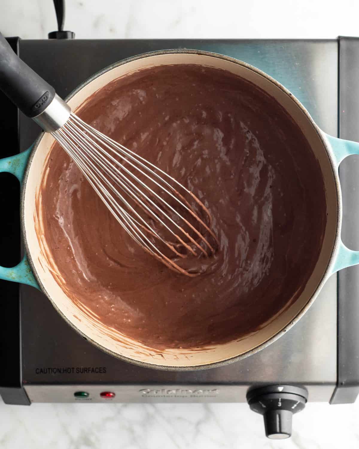 how to make chocolate ice cream - heavy cream and cocoa powder whisked together in a blue dutch oven on a metal heating plate with a metal whisk in the cream/cocoa mixture
