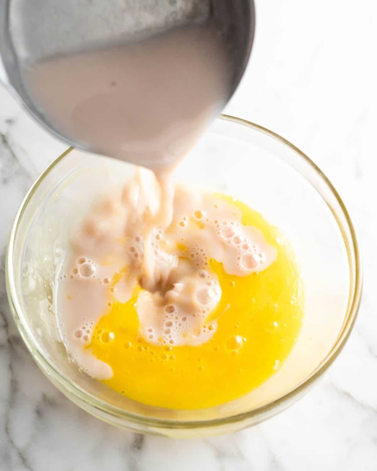 how to make chocolate ice cream - warm milk being poured into beaten egg yolks in a glass bowl on a marble surface to temper the eggs while making the custard for the chocolate ice cream