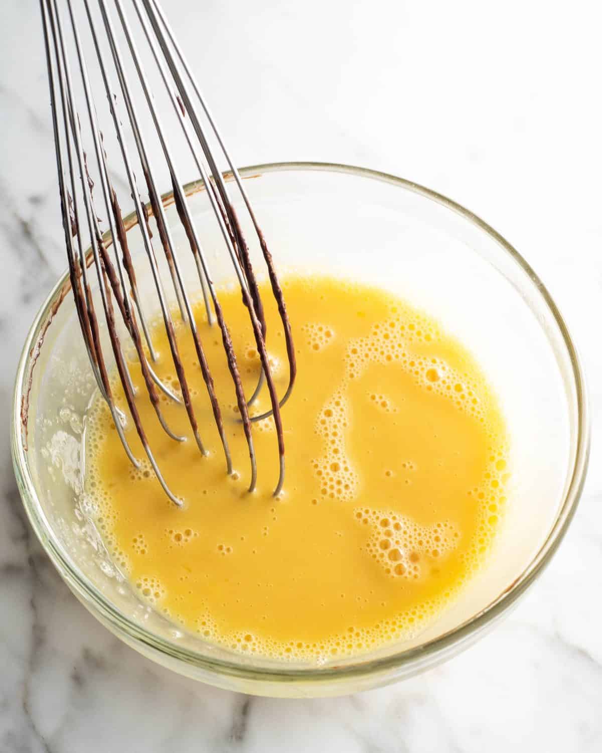 how to make chocolate ice cream - warm milk being whisked into beaten egg yolks in a glass bowl on a marble surface to temper the eggs while making the custard for the chocolate ice cream