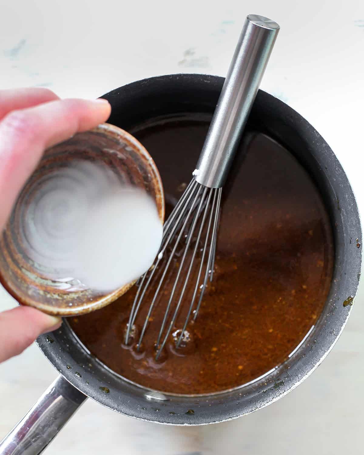 making slow cooker teriyaki chicken sauce in a pot. Pouring a cornstarch/water mixture into the sauce with a metal whisk in the pot