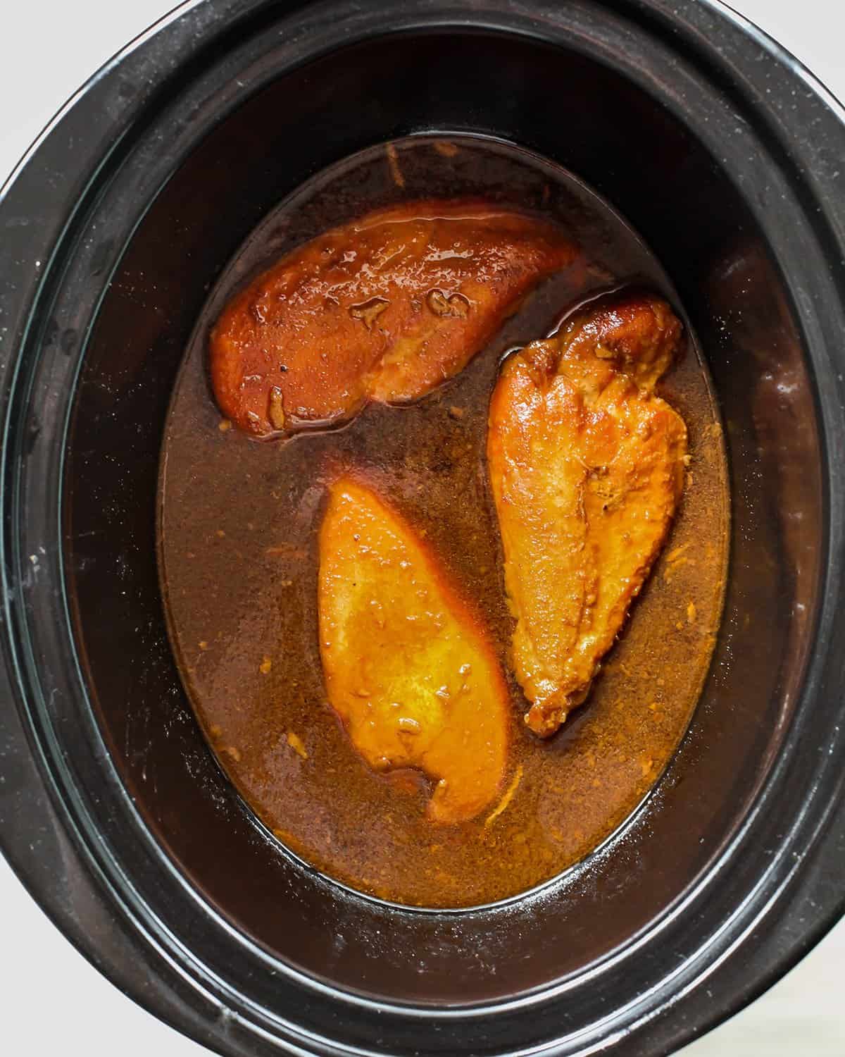 making slow cooker teriyaki chicken - three chicken breasts in the sauce in a crock pot after cooking