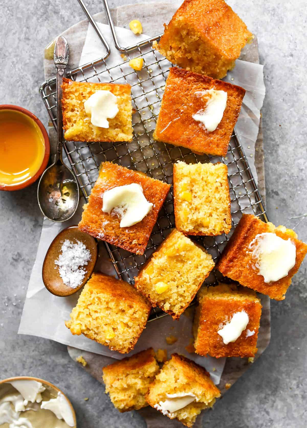 10 slices of cornbread on a wire cooling rack that's on parchment paper. Some are on their side so the moist, corn-studded texture is visible. Some have butter on top. 