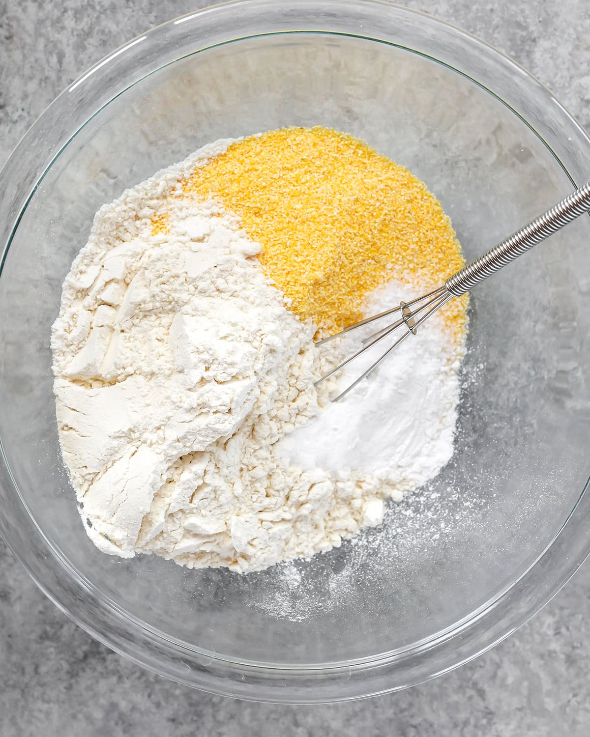 photo showing how to make cornbread - a glass bowl with flour, cornmeal, salt, baking powder and baking soda with a metal wire whisk before mixing