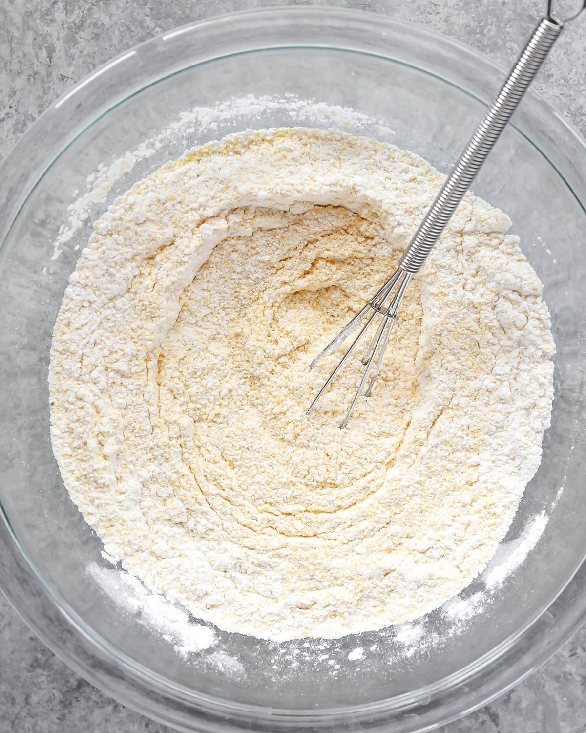 photo showing how to make cornbread - a glass bowl with flour, cornmeal, salt, baking powder and baking soda with a metal wire whisk after mixing