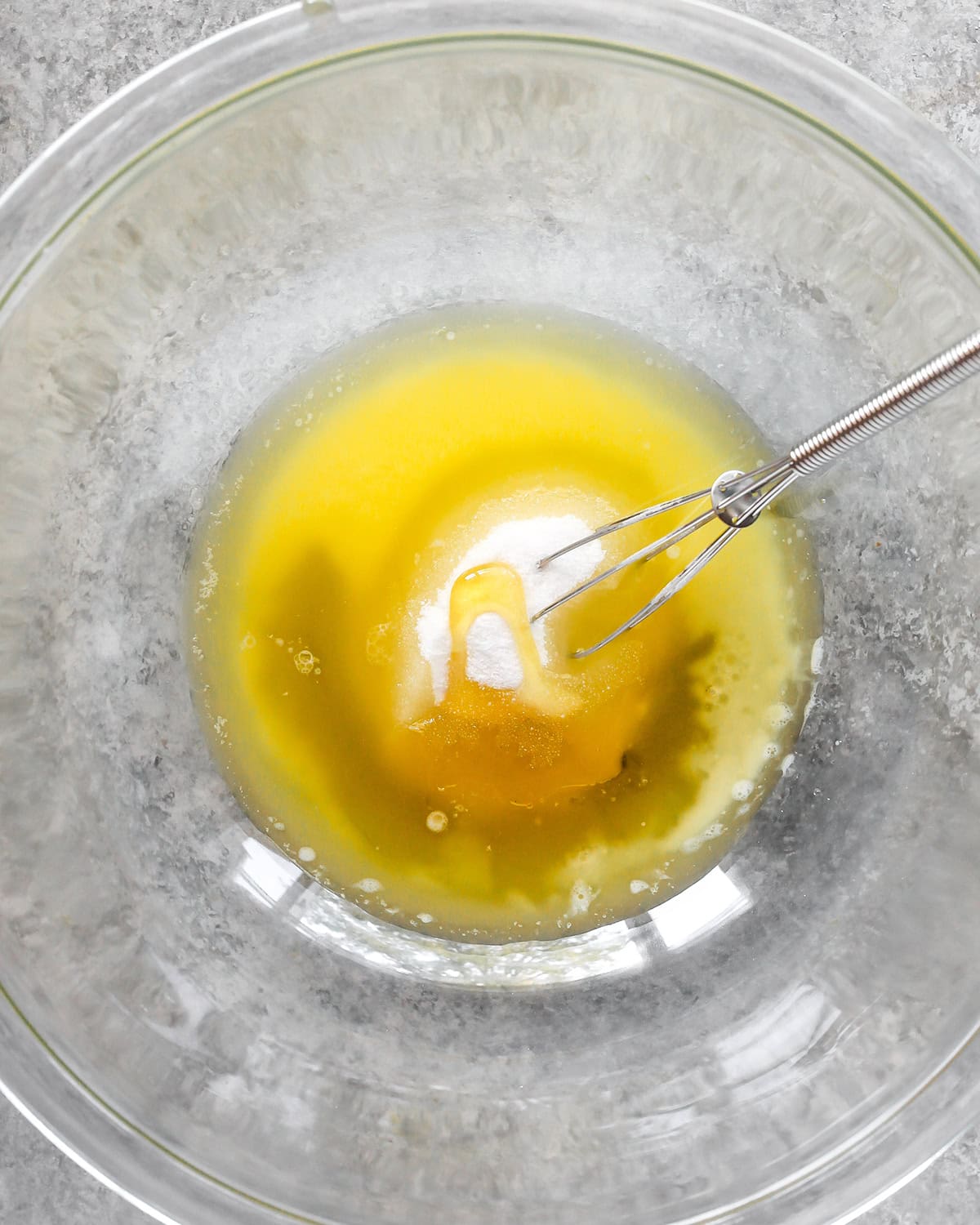 photo showing how to make cornbread -melted butter, sugar and honey in a glass bowl with a metal whisk on a gray surface before mixing