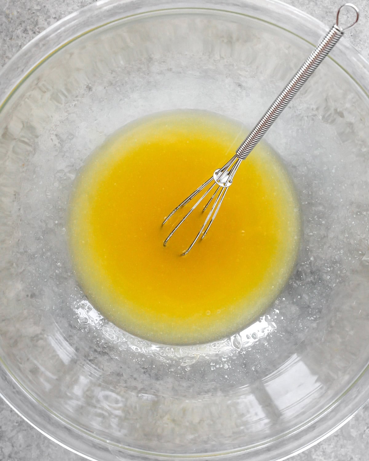 photo showing how to make cornbread -melted butter, sugar and honey in a glass bowl with a metal whisk on a gray surface after being whisked together until smooth